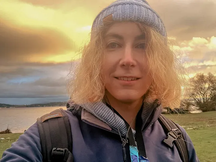 Dielle, a white transfeminine person with blue eyes, blonde hair and a blue hat, stands in front of a lake